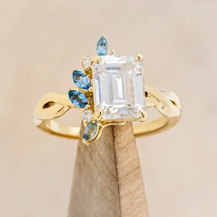 “GABRIELLA” – EMERALD CUT MOISSANITE ENGAGEMENT RING WITH AQUAMARINE & DIAMOND ACCENTS IN A TWISTED SHANK